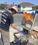 a person stands on the opposite side of the road in front of a bank of america building, painting it on a canvas. the building on the painting is burning violently. there are parodied bank logos printed on the back of the shirt the person is wearing, they are edited to say "Bad for America", "shitibank", etc